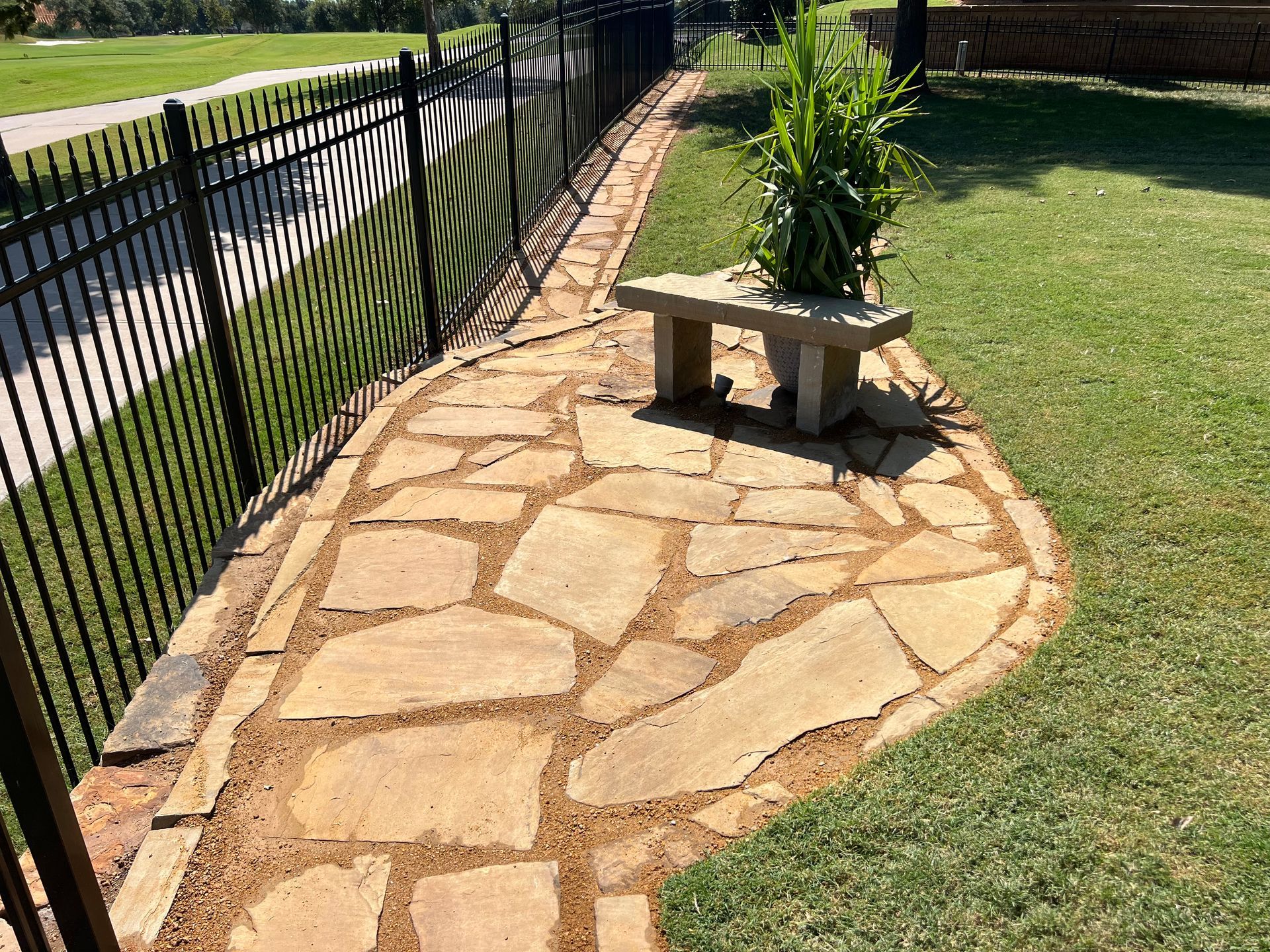 A stone pathway runs alongside a black metal fence, leading to a small concrete bench with a potted plant on it.