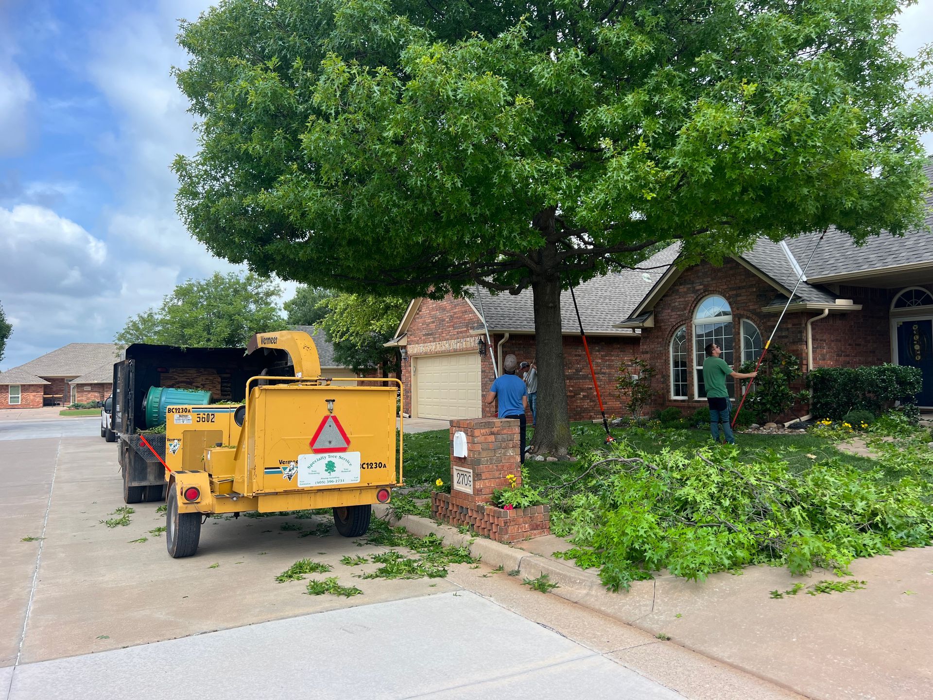 Two workers trim a tree in front of a brick house while a yellow wood chipper sits on the driveway nearby.