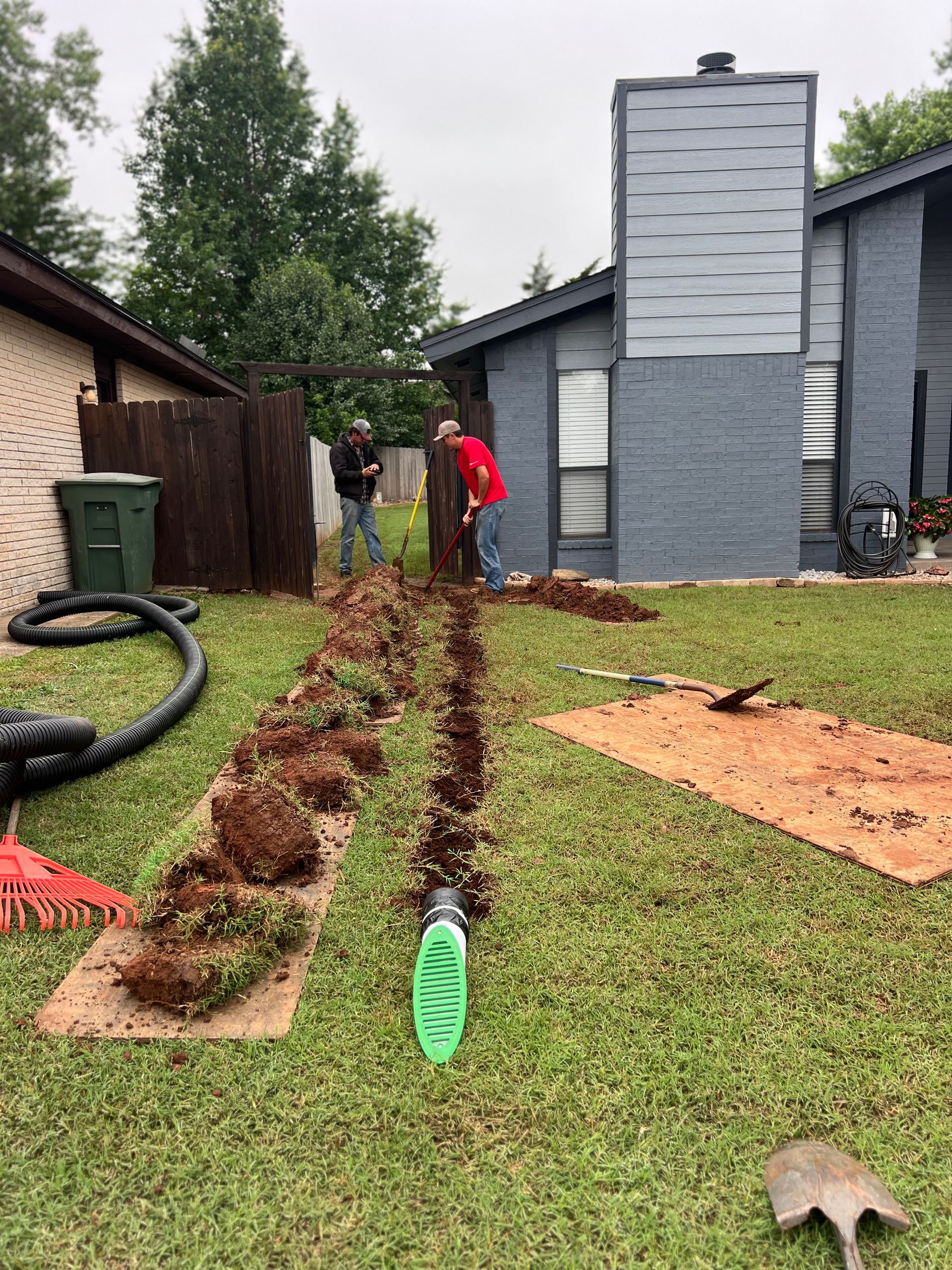 Two people dig trenches in a residential lawn near a house, with a green drainage grate placed in the foreground.