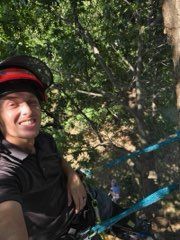A person wearing a climbing helmet and safety harness smiles while suspended in a tree canopy.