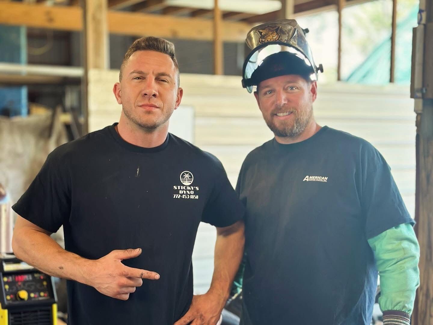 Two men are standing next to each other and one is wearing a welding helmet.