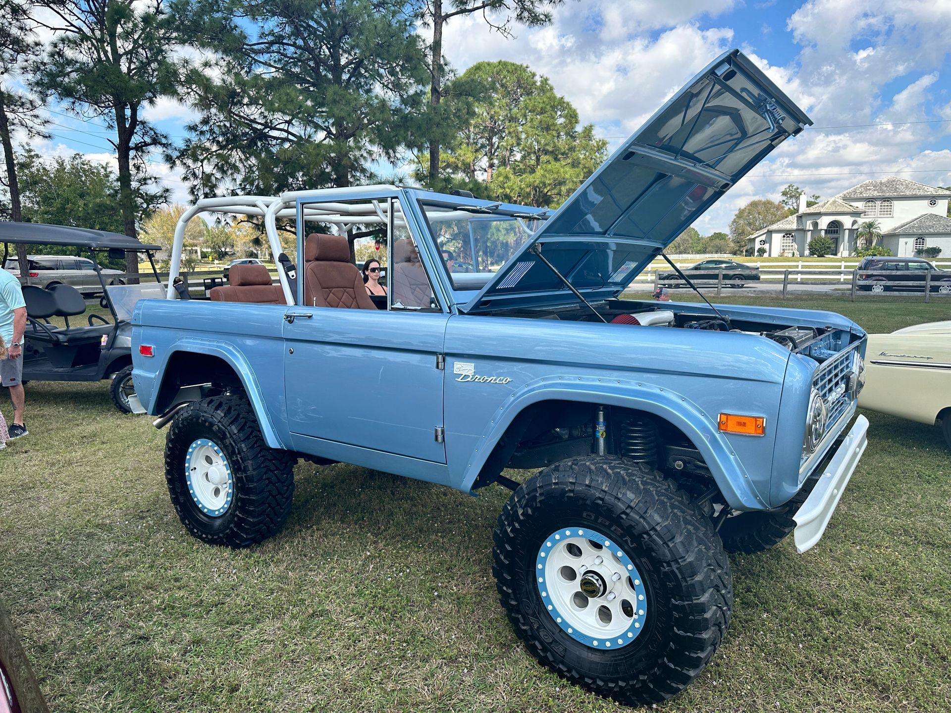 A blue ford bronco with the hood up is parked in the grass at a car show.