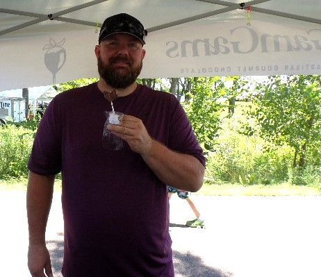 GamGams chocolate vendor tent. He wears a purple shirt and cap, smiles outside on a sunny day.