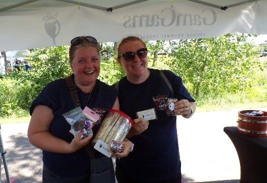 Two women smiling, holding packaged snacks at GamGams outdoor market.