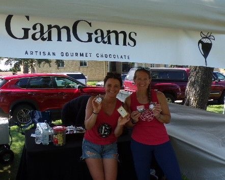 Two women at a GamGams Artisan Gourmet Chocolate booth. They hold chocolate, wearing red shirts, and smiling.