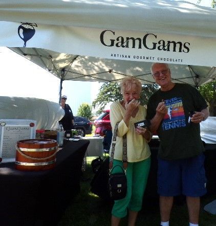 Couple at a chocolate stand: GamGams, smiling while tasting chocolate, sunny outdoor setting.