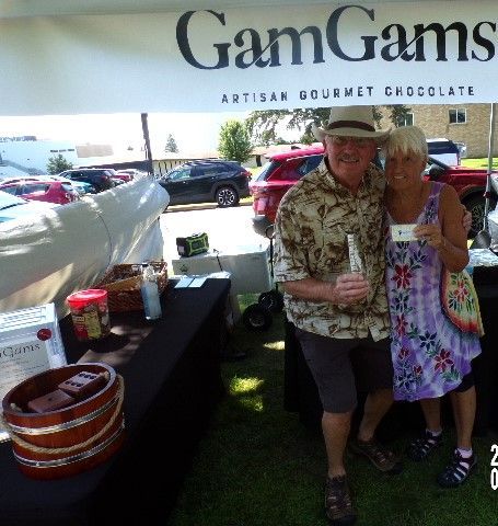 Couple at GamGams Artisan Gourmet Chocolate stand. Man holds bottle, woman displays product. Outdoor setting, sunny.