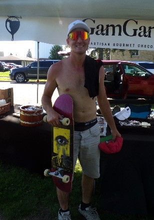 Man, shirtless, holding skateboard, smiling, stands at GamGar artisan booth.