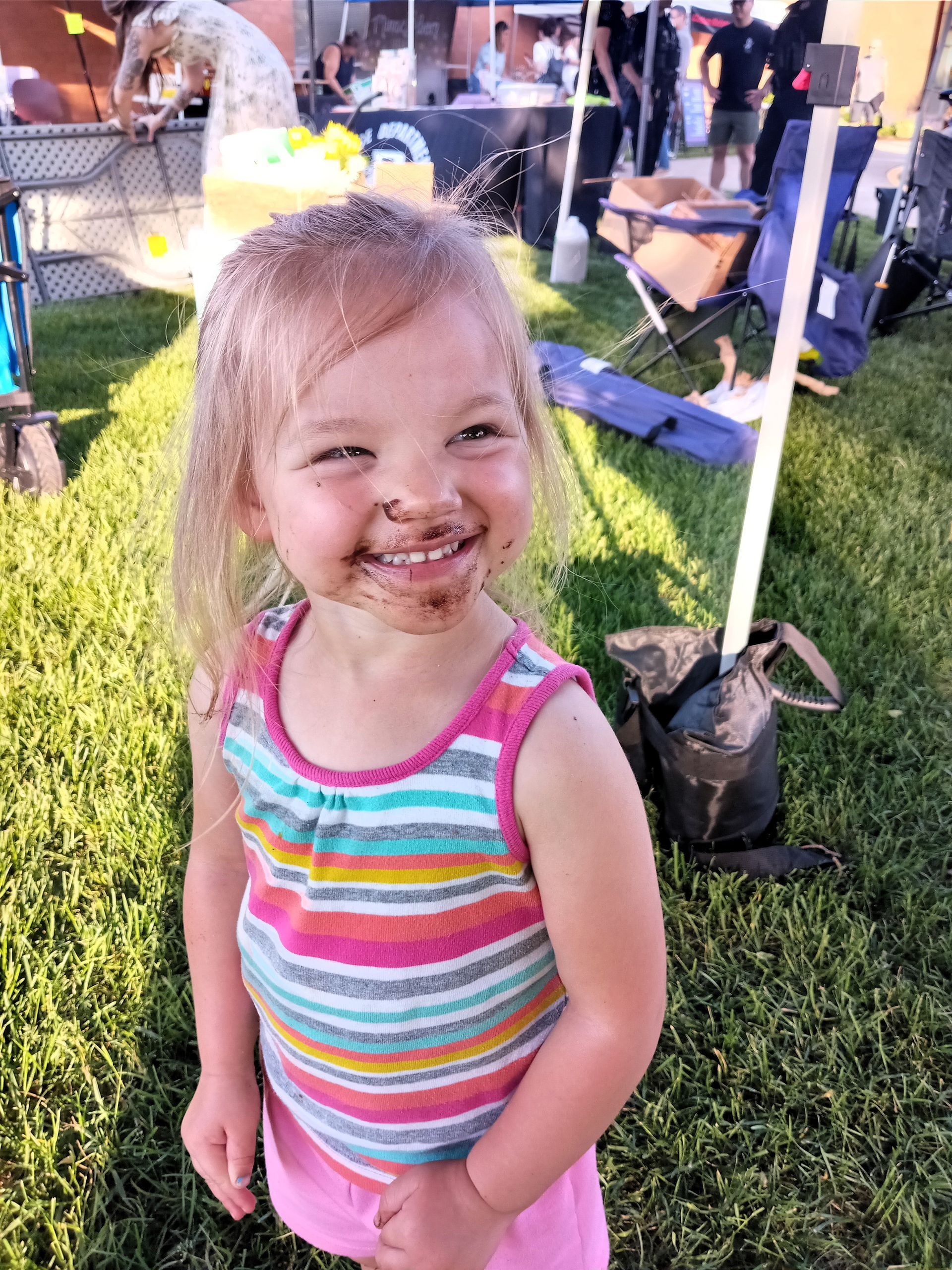 Smiling neice in a rainbow striped tank top standing on grass at an outdoor event with vendor tents.