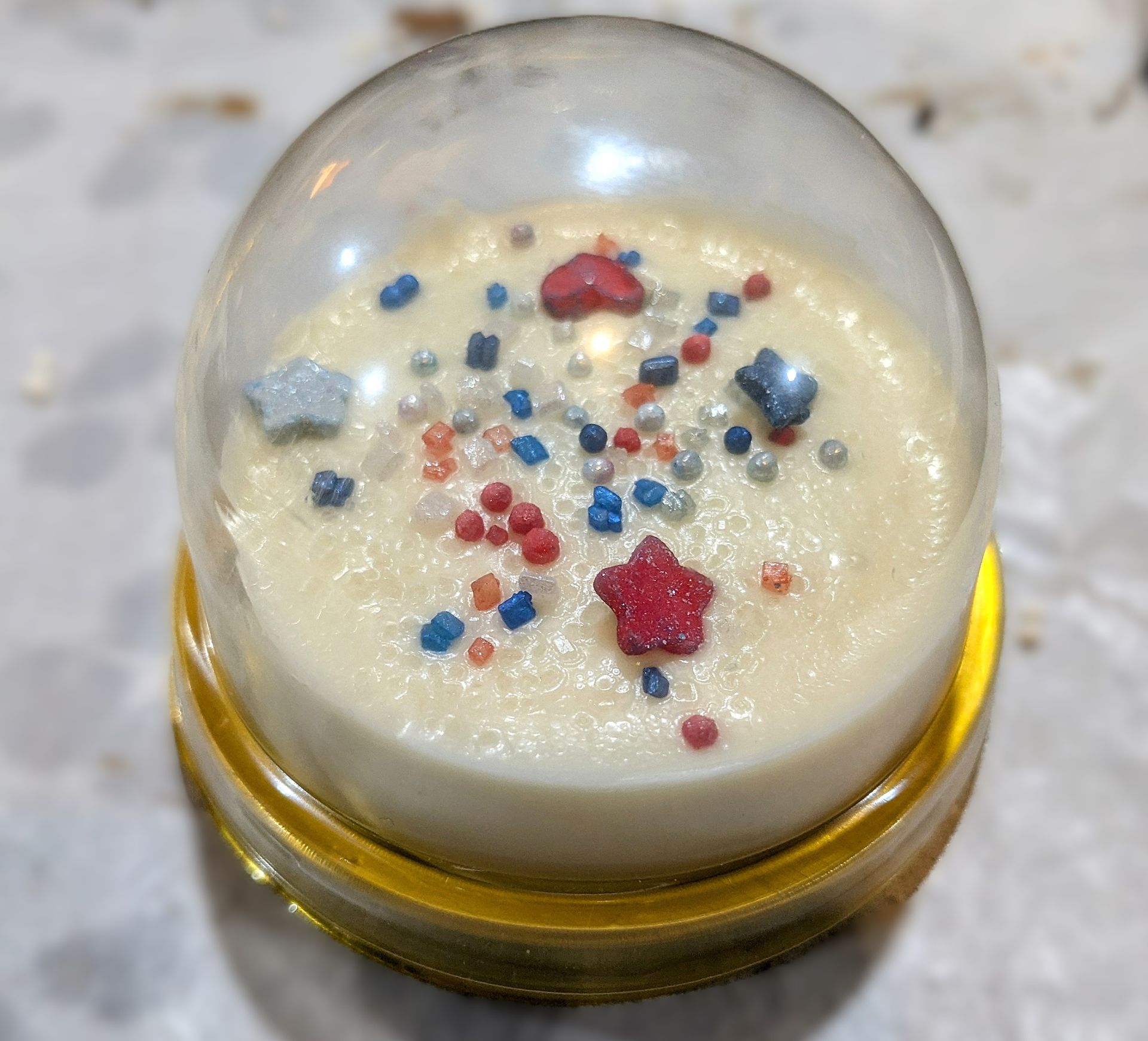 A small, white dessert topped with red, white, and blue sprinkles and stars, inside a clear dome.