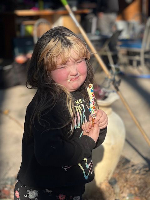 Young girl with blonde-streaked hair, holding a lollipop, from GamGams Chocolate Booth. Smiles outdoors in sunlight.