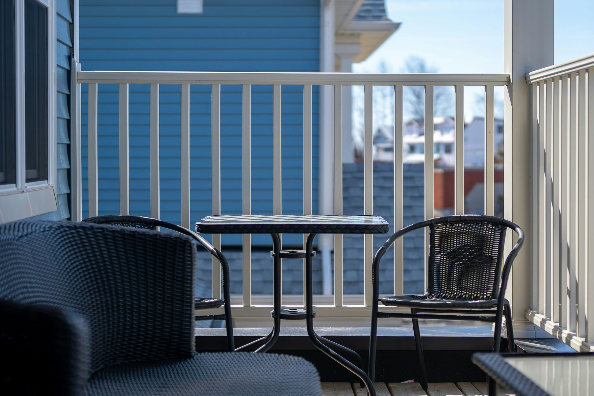 A small, dark woven patio table and chairs sit on a deck with white railings against a blue-sided house.