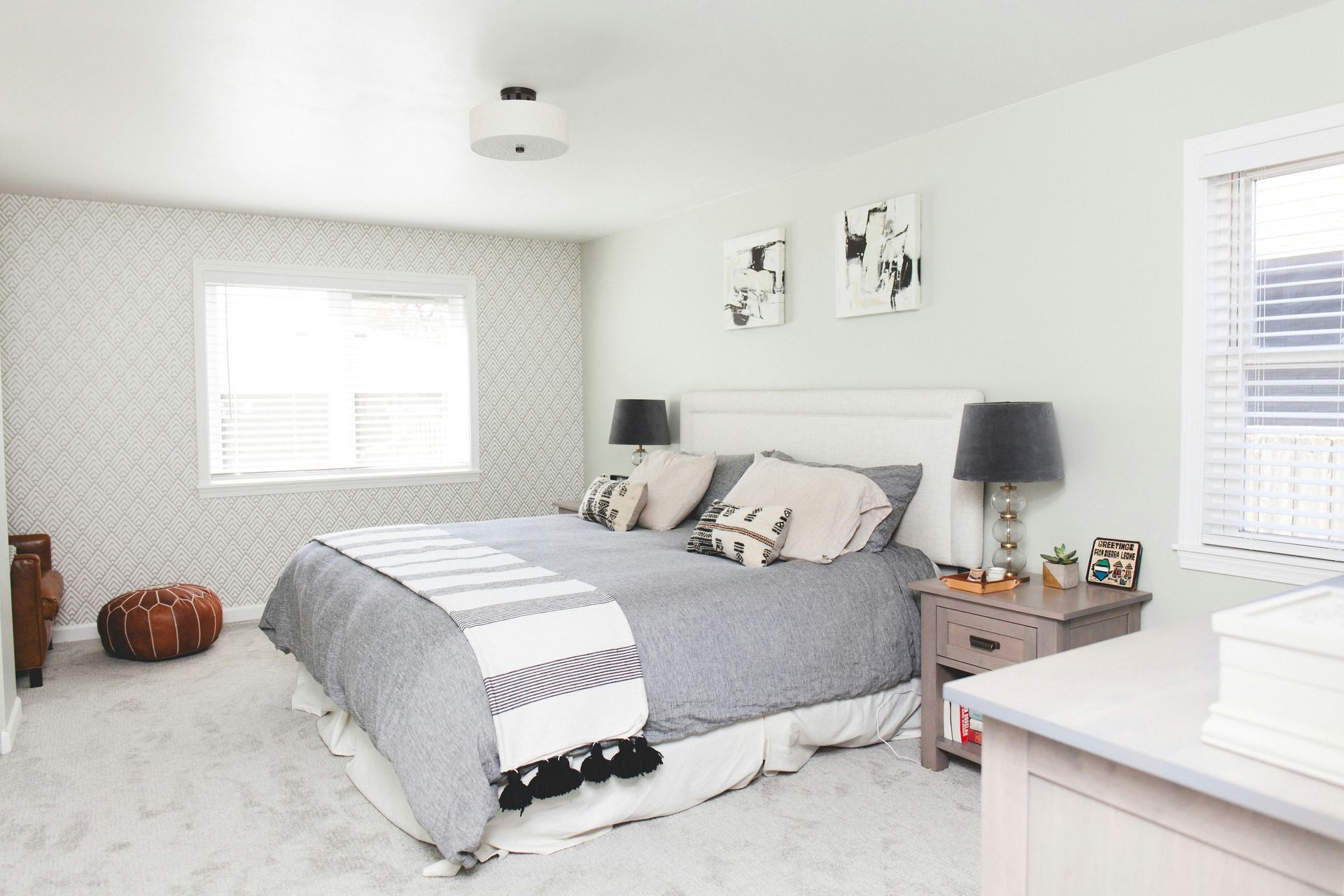 A bright bedroom with a king-sized bed, grey patterned bedding, a textured rug, two lamps, and patterned wallpaper.