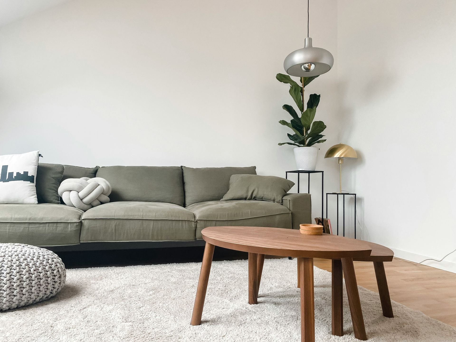A modern living room with an olive green sofa, wooden coffee table, grey floor cushion, and a hanging silver lamp.