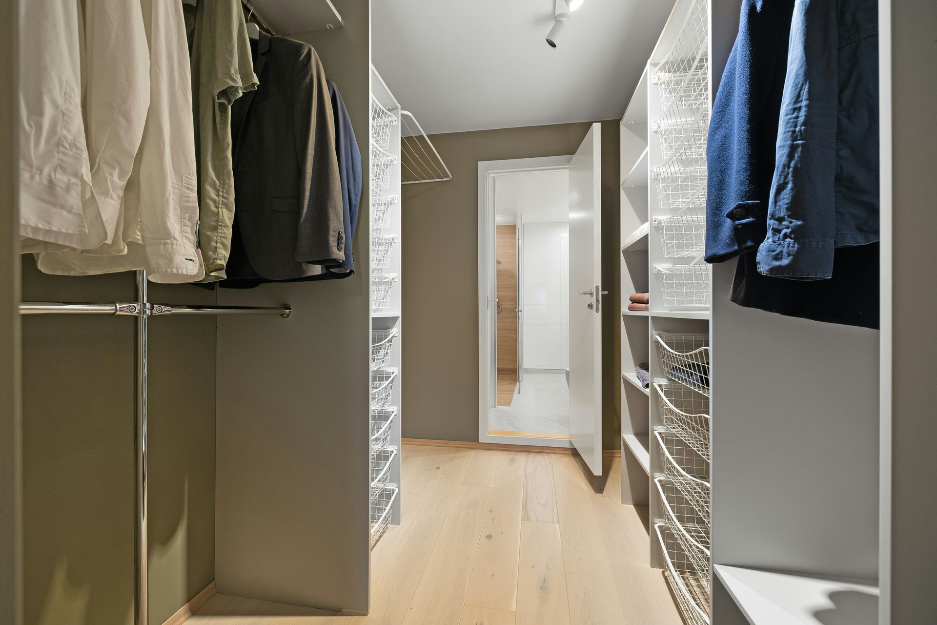 A walk-in closet with hanging clothes on both sides, white wire shelving units, and a doorway leading to another room.