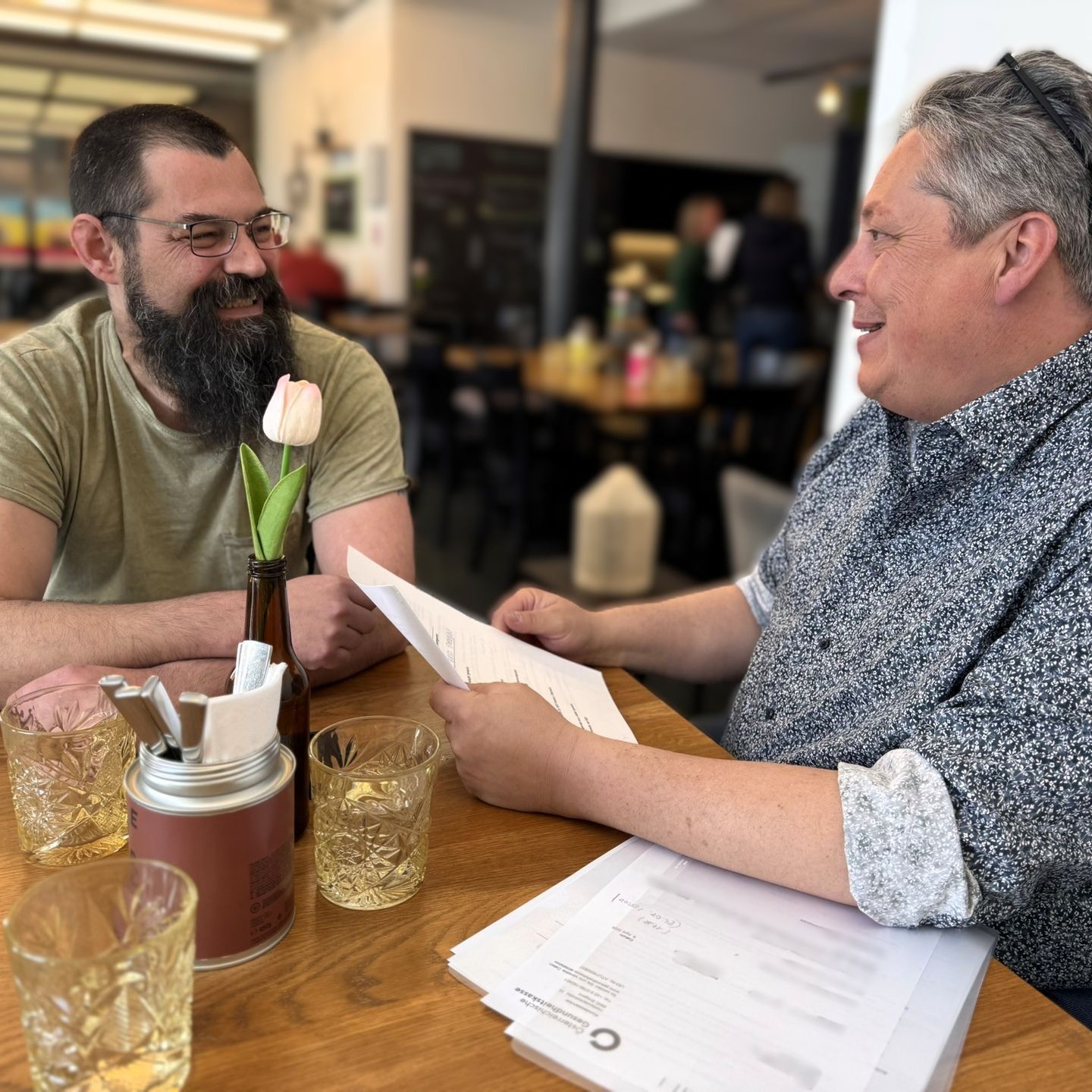 Zwei Männer sitzen an einem Cafétisch, einer hält Papiere in der Hand und spricht, der andere lächelt und hat einen Bart.