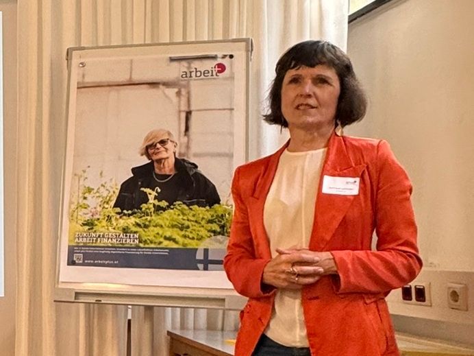 Eine Frau in einer orangefarbenen Jacke steht vor einem Plakat.