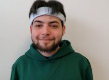 Ein junger Mann mit Bart trägt ein Stirnband und einen grünen Kapuzenpullover.