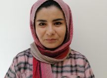 Eine Frau mit Hijab und kariertem Hemd steht vor einer weißen Wand.