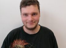 Ein junger Mann mit Bart trägt ein schwarzes T-Shirt und lächelt.