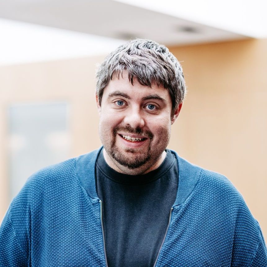 Ein Mann mit Bart trägt einen blauen Pullover und ein schwarzes Hemd.