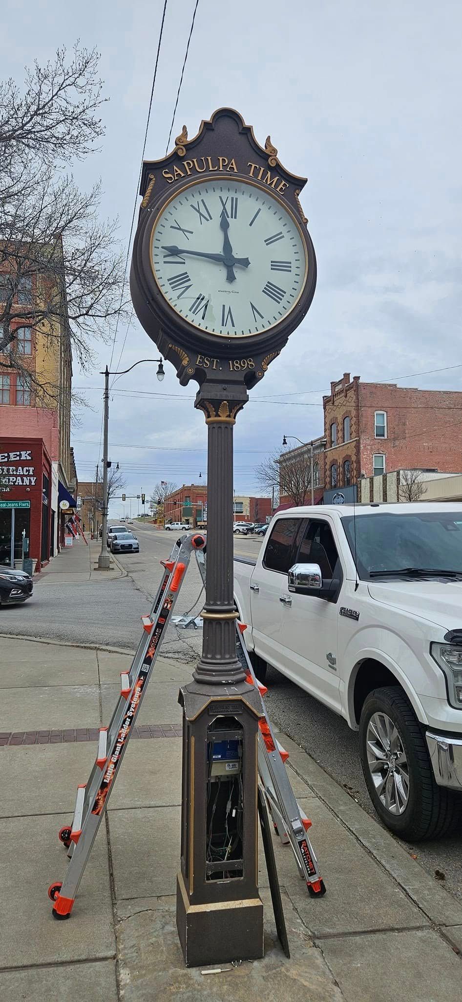 A clock is sitting on the side of a street next to a white truck.