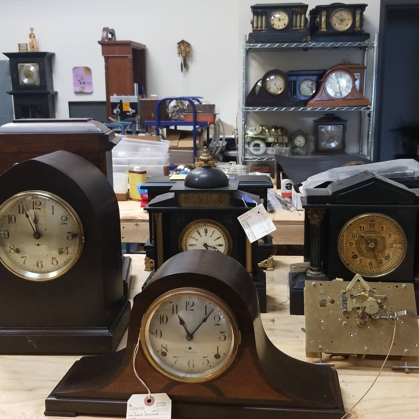 A group of clocks are sitting on a table and one of them has a tag that says ' antique ' on it