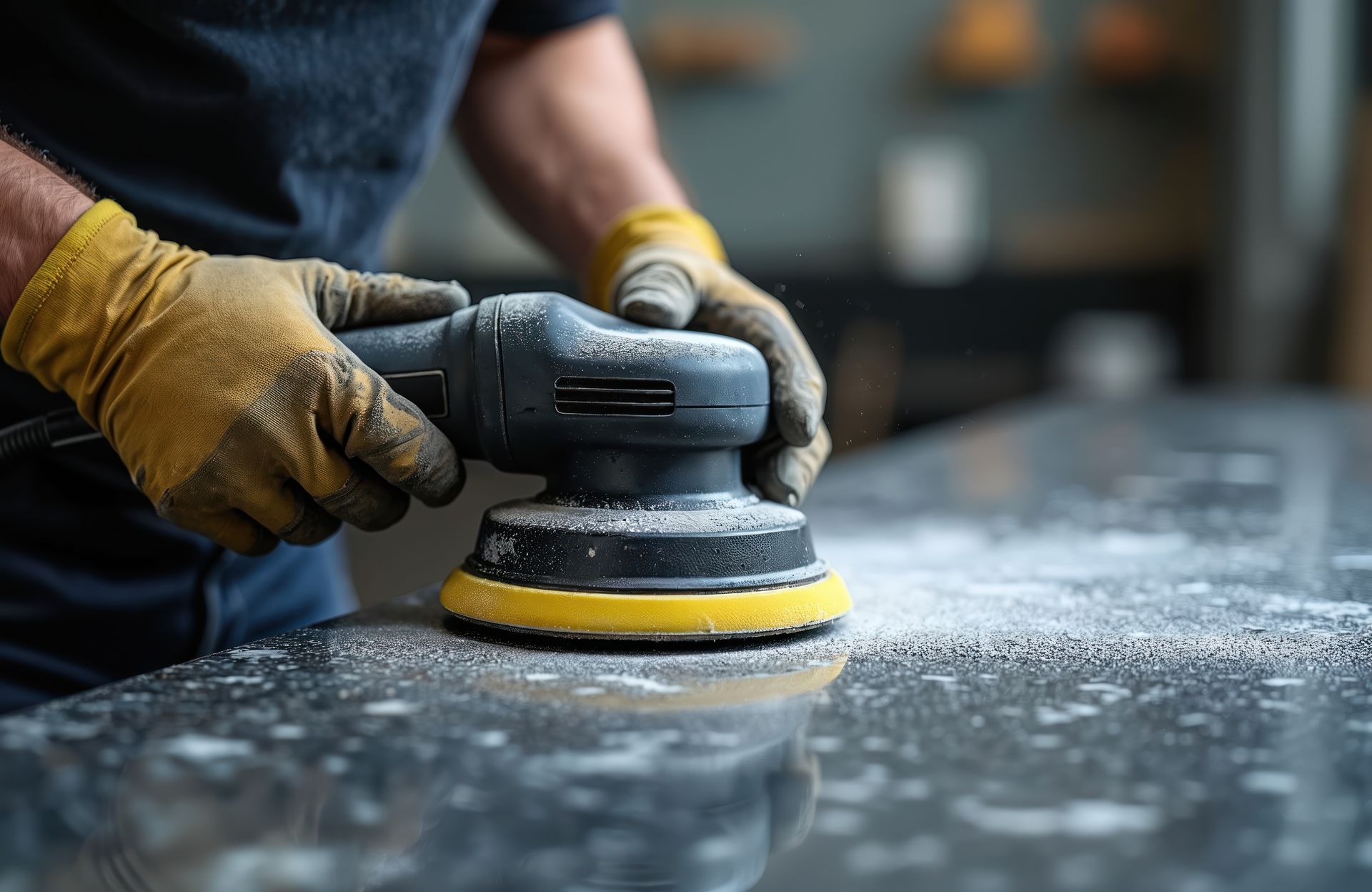 Person wearing gloves sanding a dark surface with an electric sander, creating dust.