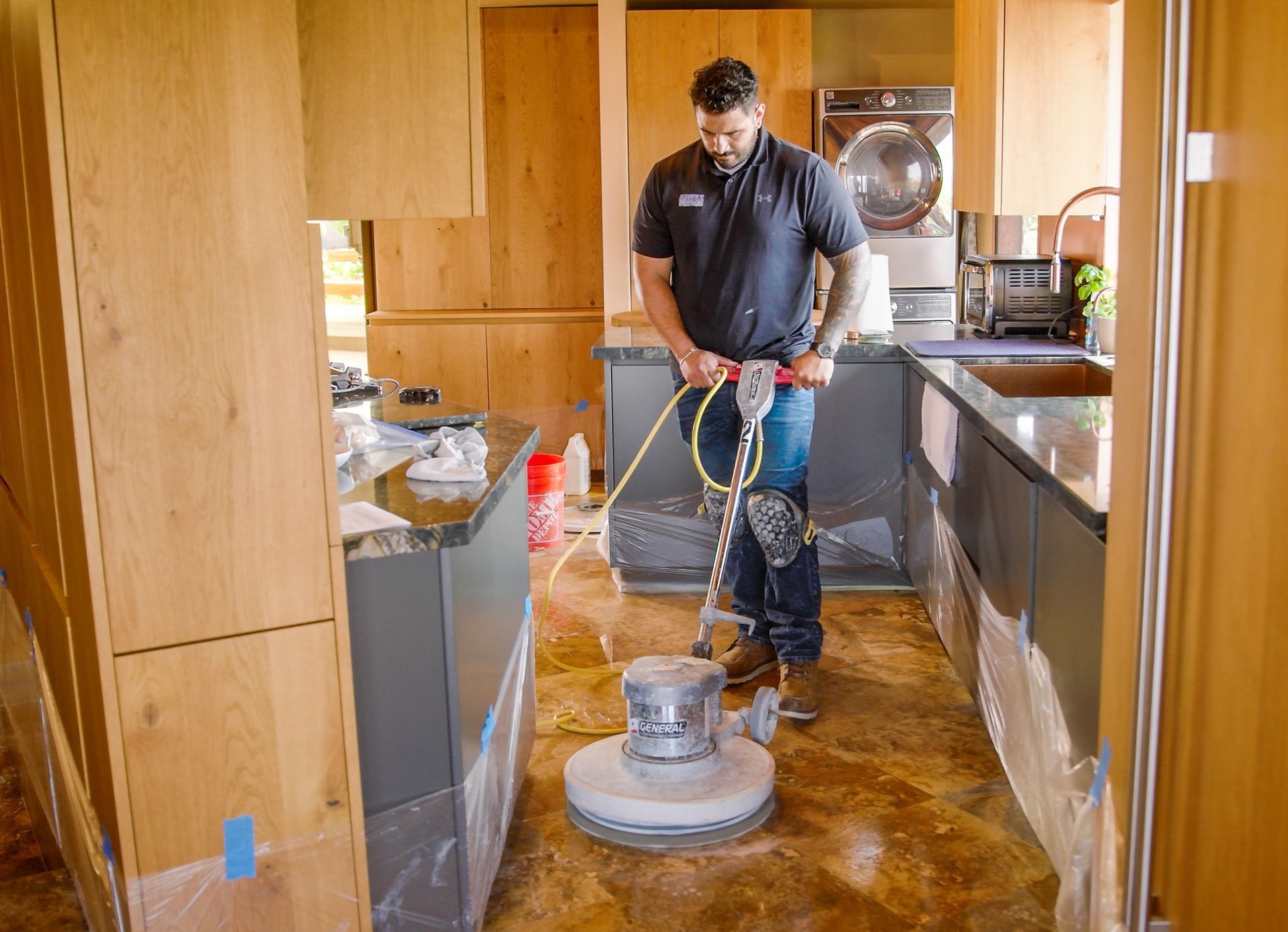 A worker in a dark shirt uses a floor buffer on a brown tiled floor inside a modern kitchen with wood cabinets.