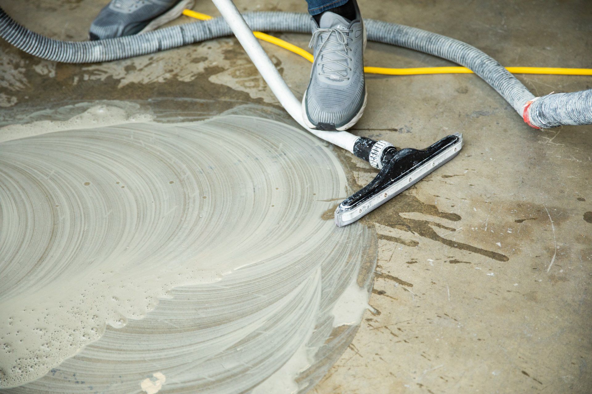 Person using a wet vacuum to clean a soapy concrete floor; white hose and nozzle.