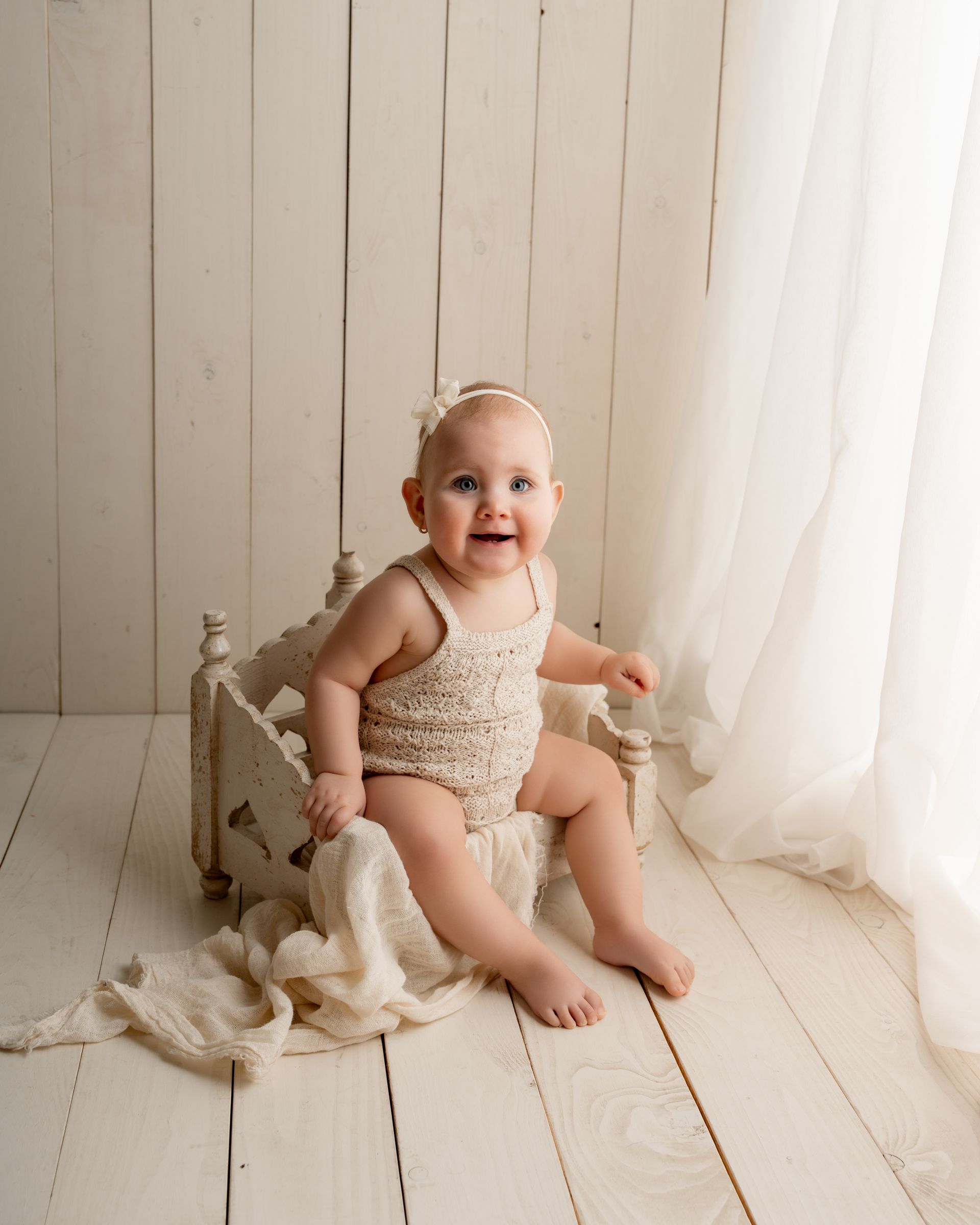 Een baby ligt lachend in een vintage bedje, gekleed in een gebreid rompertje en hoofbandje, met een dekentje eroverheen, in een lichte omgeving.