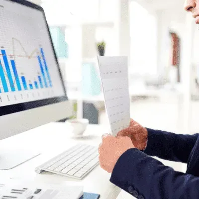 A man is sitting at a desk in front of a computer holding a piece of paper.