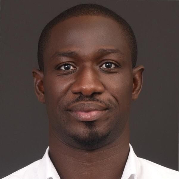 Close-up of a Black man with short hair, smiling slightly. Wearing a white shirt, against a gray background.
