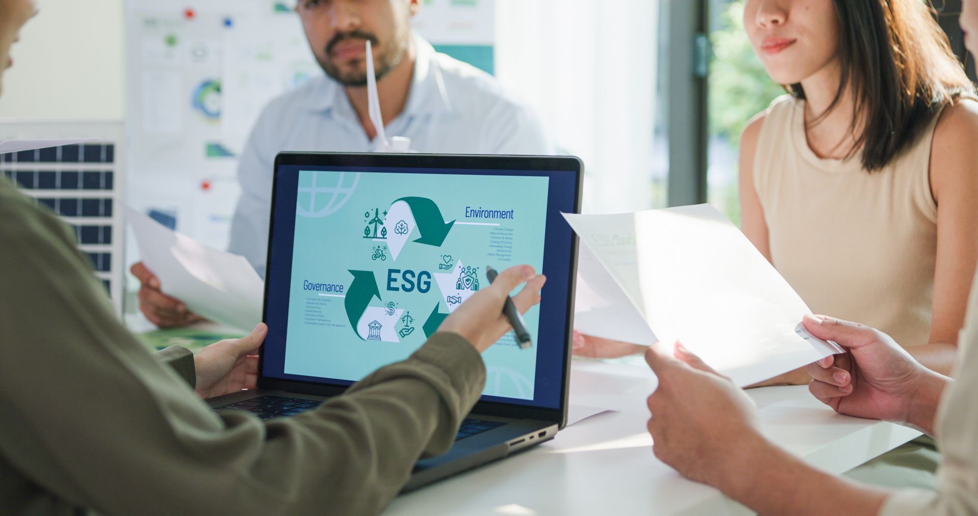 People reviewing ESG data on a laptop screen, discussing and holding papers in an office setting.