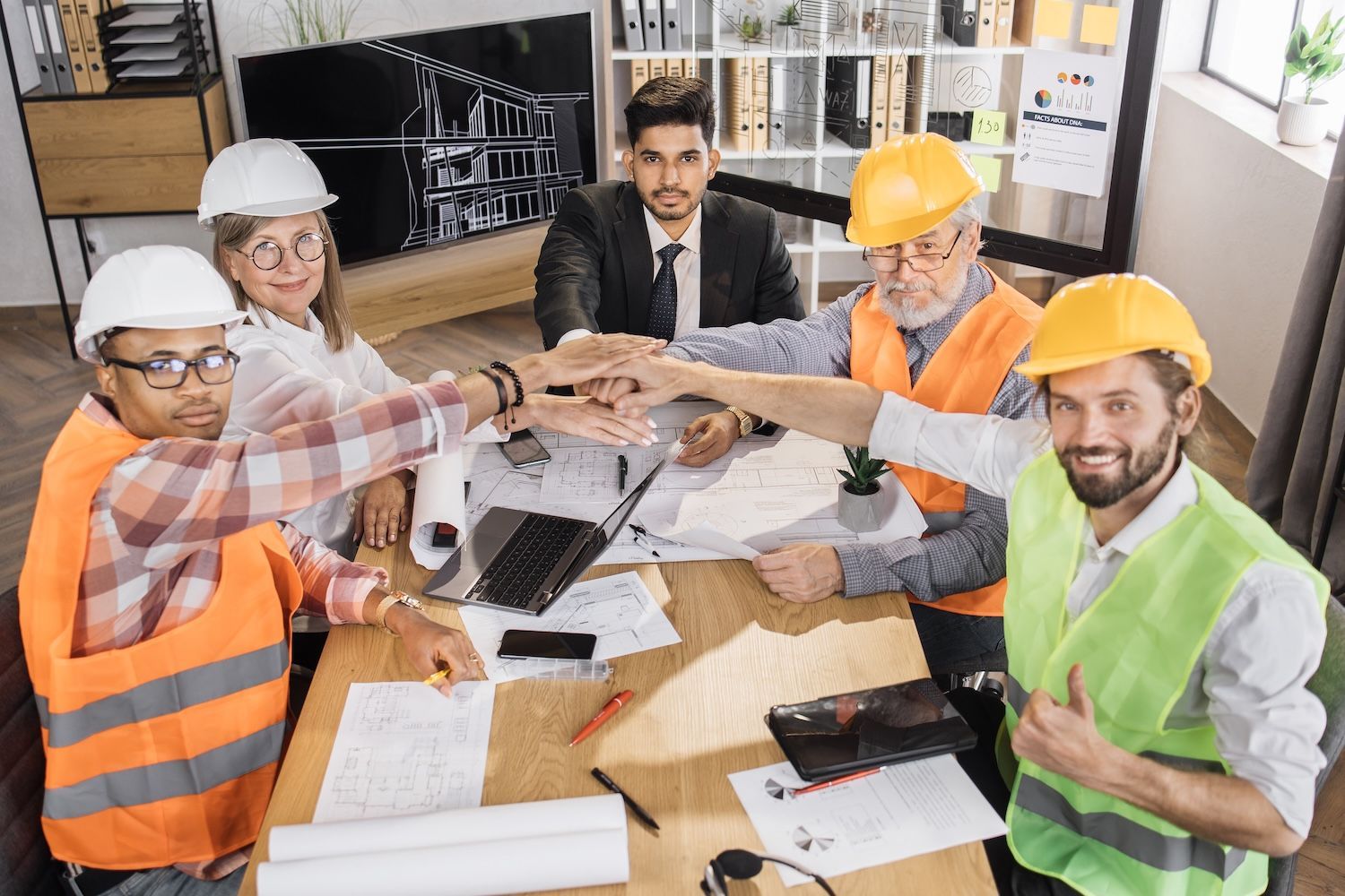 Team of construction workers and a business professional celebrate with hands stacked together at a table, smiling.