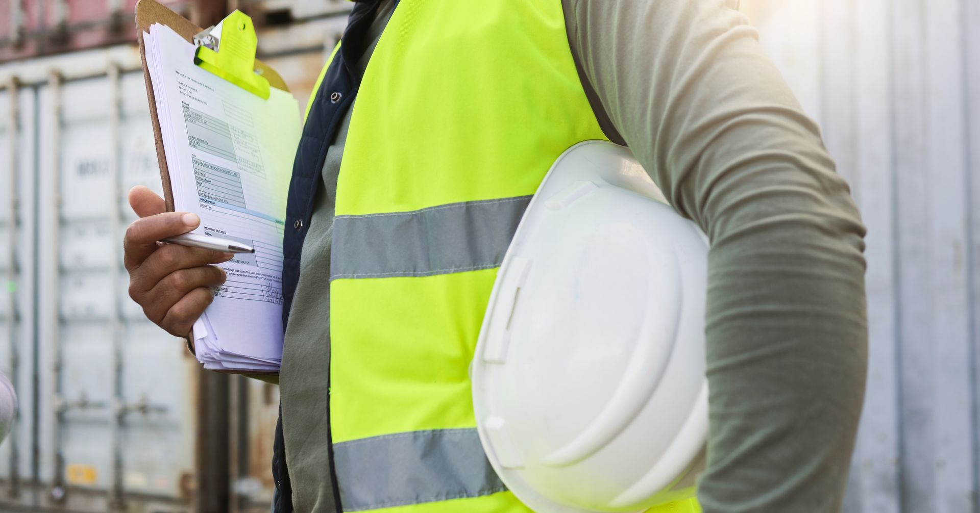 Person in safety vest holding clipboard and hard hat next to cargo containers.