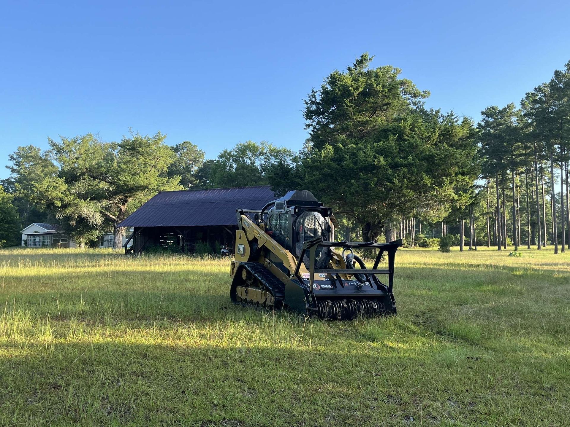 A small, green and gold machine in a grassy field, near a dark-roofed building and trees under a blue sky.
