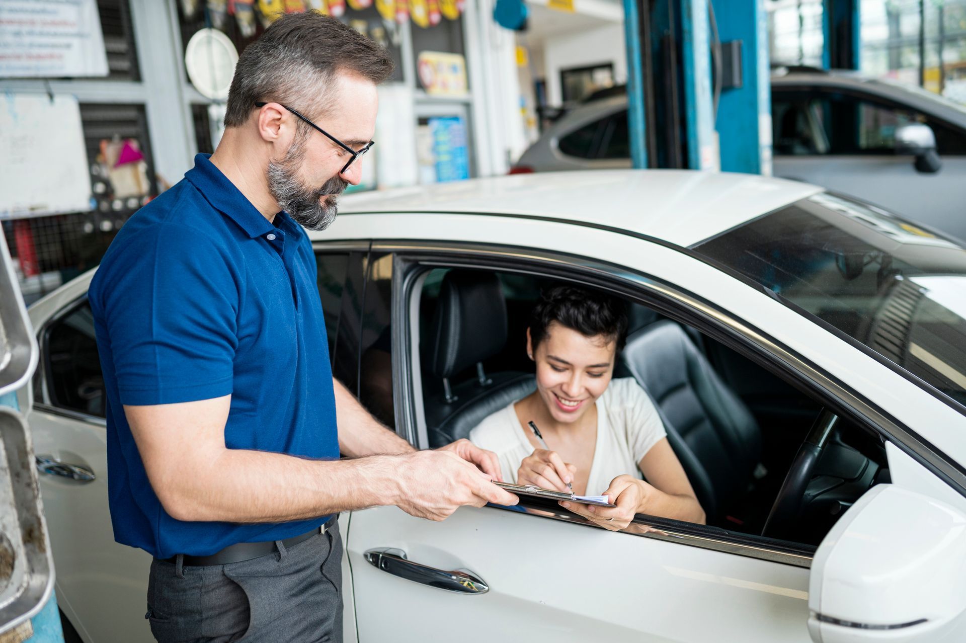 Mechanic handing paperwork to a driver in a white car at a repair shop; the driver is signing.