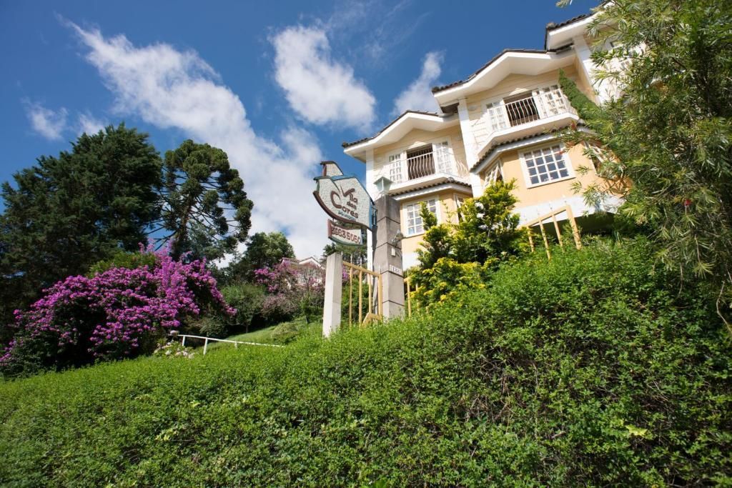 Um prédio de vários andares, cor creme, situado em uma encosta gramada sob um céu azul, com uma placa e flores roxas nas proximidades.