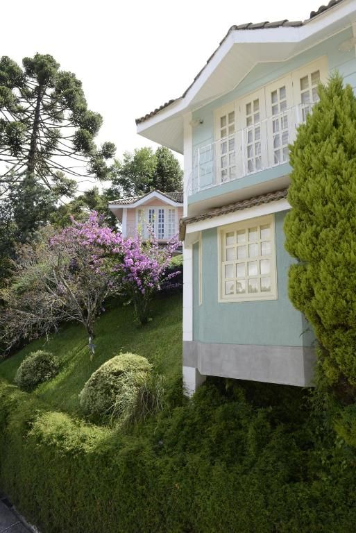 Uma casa de dois andares, azul-clara com detalhes em branco, situada em uma colina gramada ao lado de uma árvore florida e uma grande conífera.