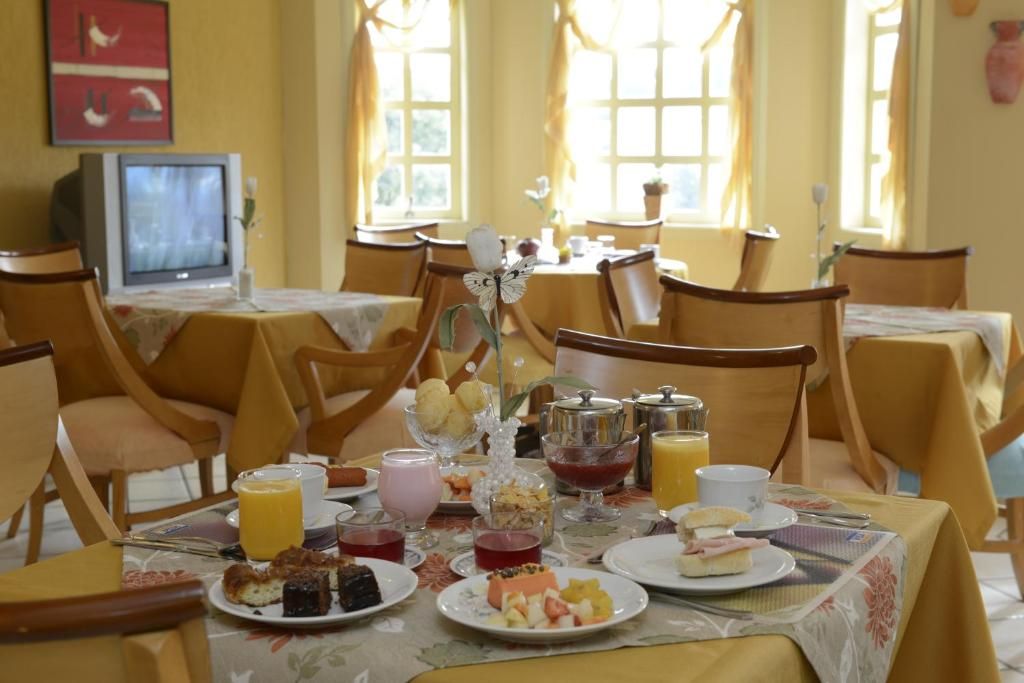 Uma mesa de café da manhã posta com doces, frutas e bebidas em uma sala de jantar ensolarada, com tons de amarelo.