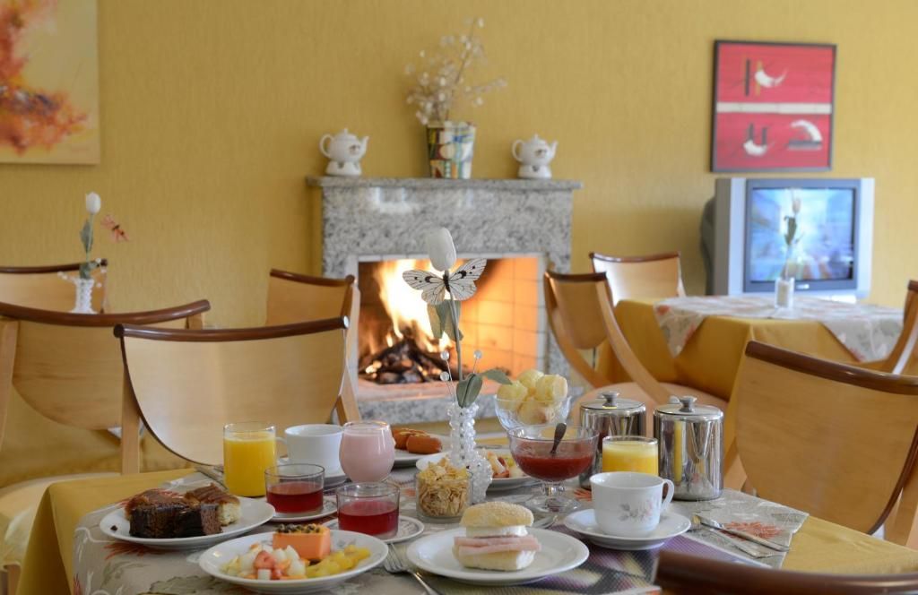 Uma mesa posta para o café da manhã em uma sala aconchegante e ensolarada, com uma lareira acolhedora, cadeiras de madeira clara e paredes amarelas.