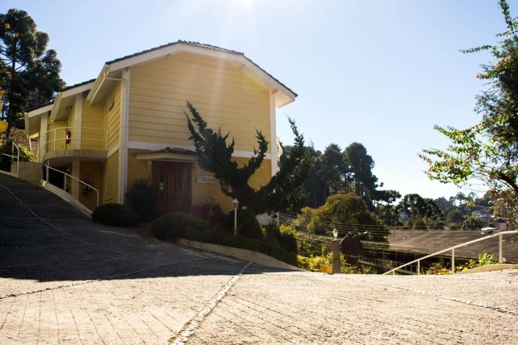 Uma casa amarela de dois andares se ergue no topo de uma entrada de automóveis pavimentada com pedras, sob um céu claro e ensolarado.