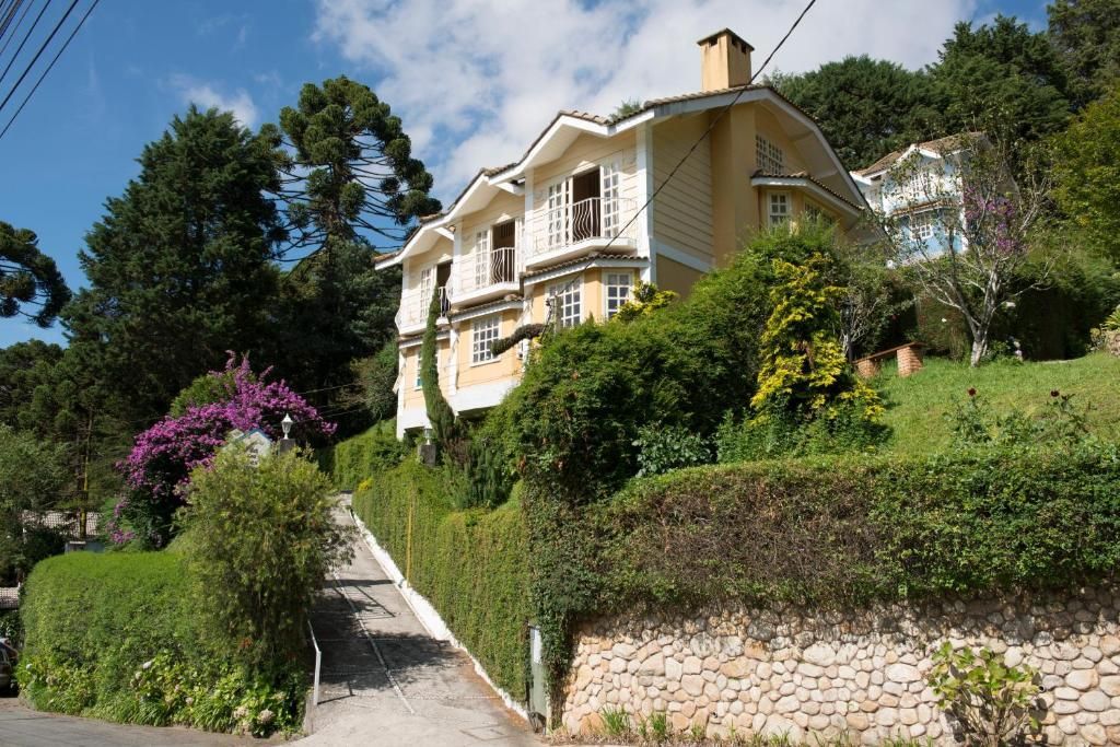 Uma casa de vários andares, de cor creme, está situada em uma encosta verdejante e exuberante, com um muro de contenção de pedra ao longo de um caminho pavimentado.