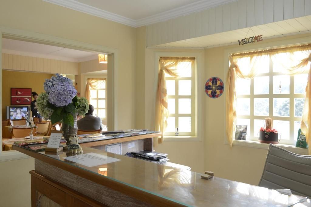 Uma recepção em uma sala bem iluminada, de cor creme, com duas janelas, cortinas amarelas translúcidas e um buquê de flores.