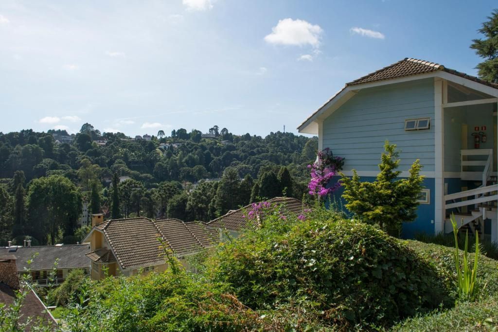 Uma casa azul-clara se ergue em uma colina com vista para florestas verdejantes e vinhedos em terraços, sob um céu azul brilhante.