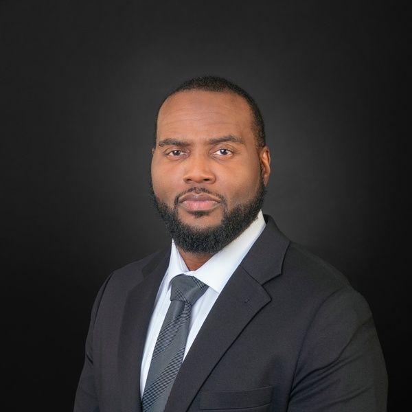 Professional headshot of a man in a black suit and gray tie against a dark background