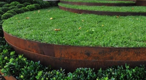 Curved, weathered steel retaining walls create terraced grass levels in a landscaped garden with low green shrubs. — BVN Landscape & Steel Supplies in St Georges Basin, NSW