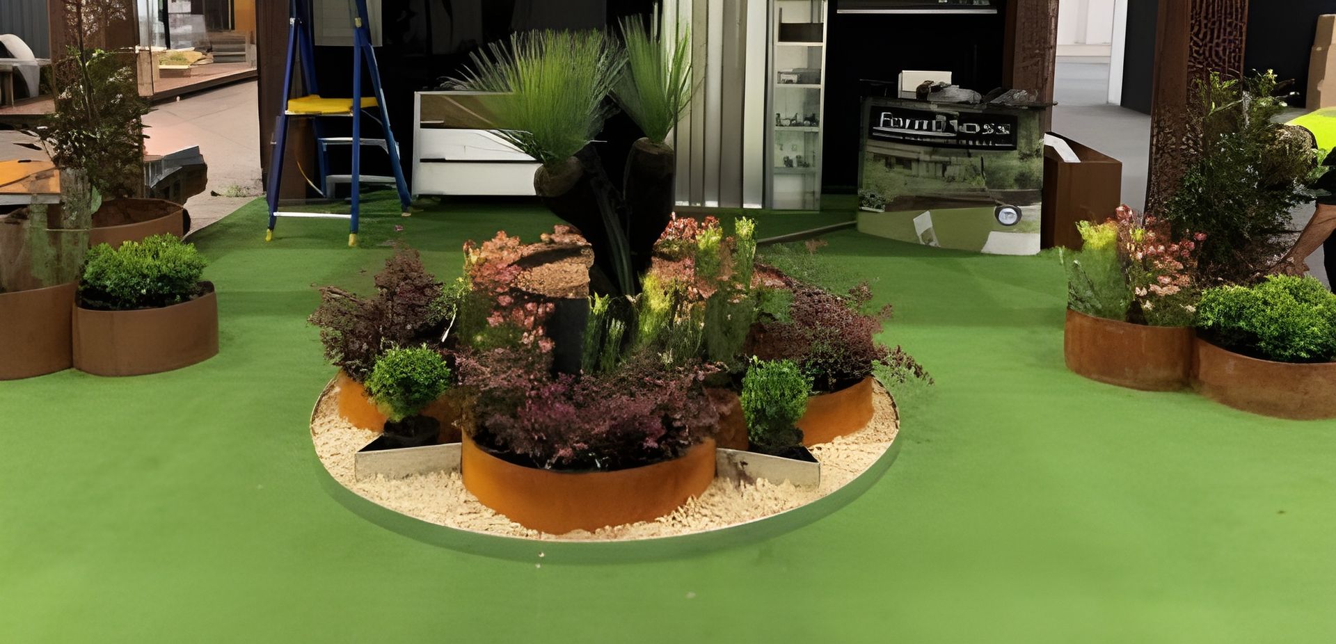 A circular indoor planter display featuring various green shrubs and grasses set in rust-coloured containers on a green floor. — BVN Landscape & Steel Supplies in Nowra, NSW