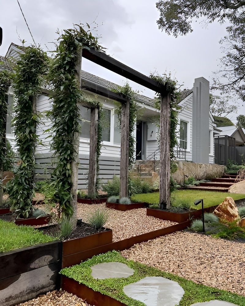 A modern front garden with a rusted metal pergola, climbing vines, stone stepping stones, and gravel pathways. — BVN Landscape & Steel Supplies in St Georges Basin, NSW